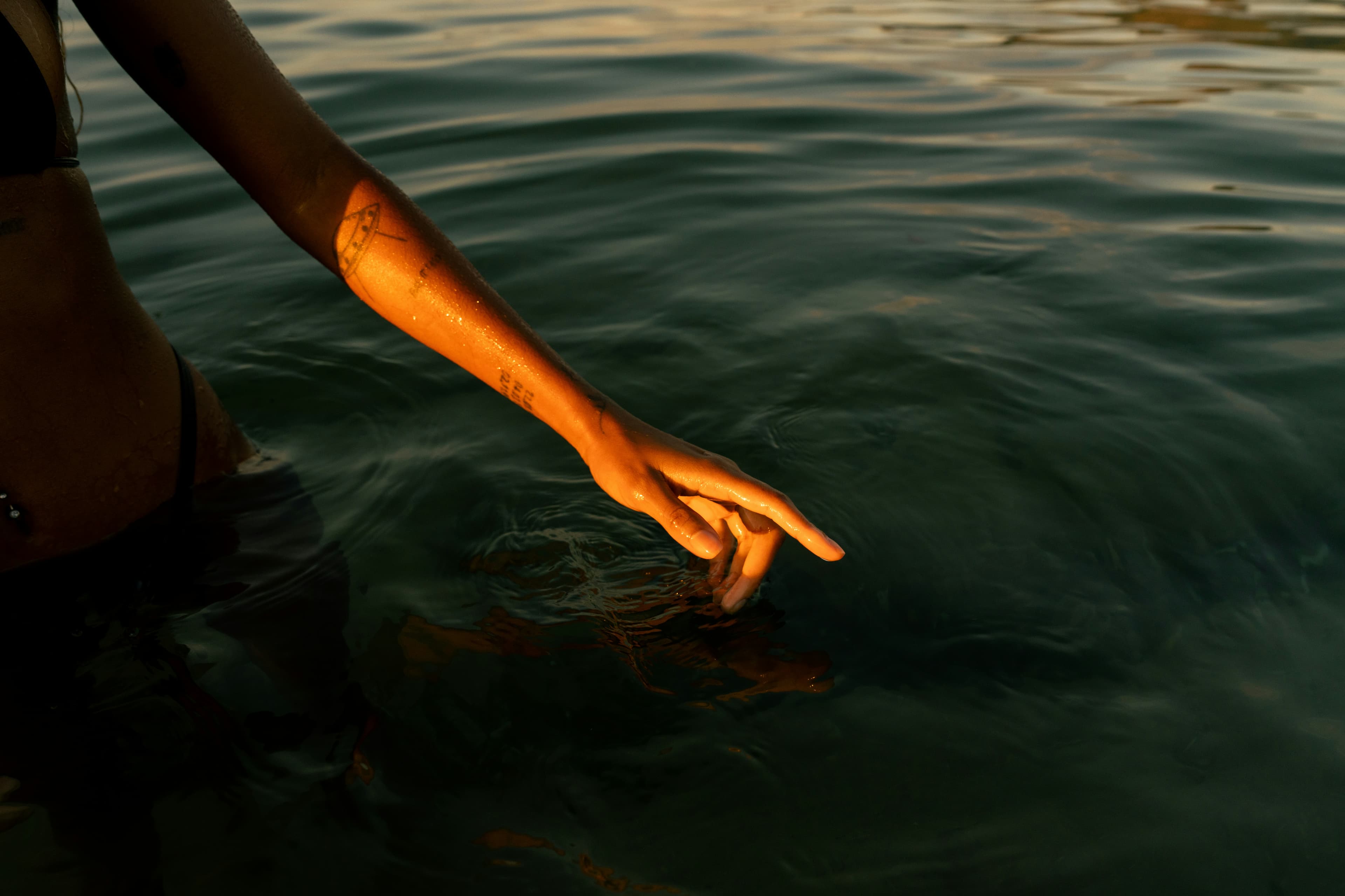 A person's arm partially submerged in water, with sunlight casting a warm glow on the skin and creating ripples in the dark water.