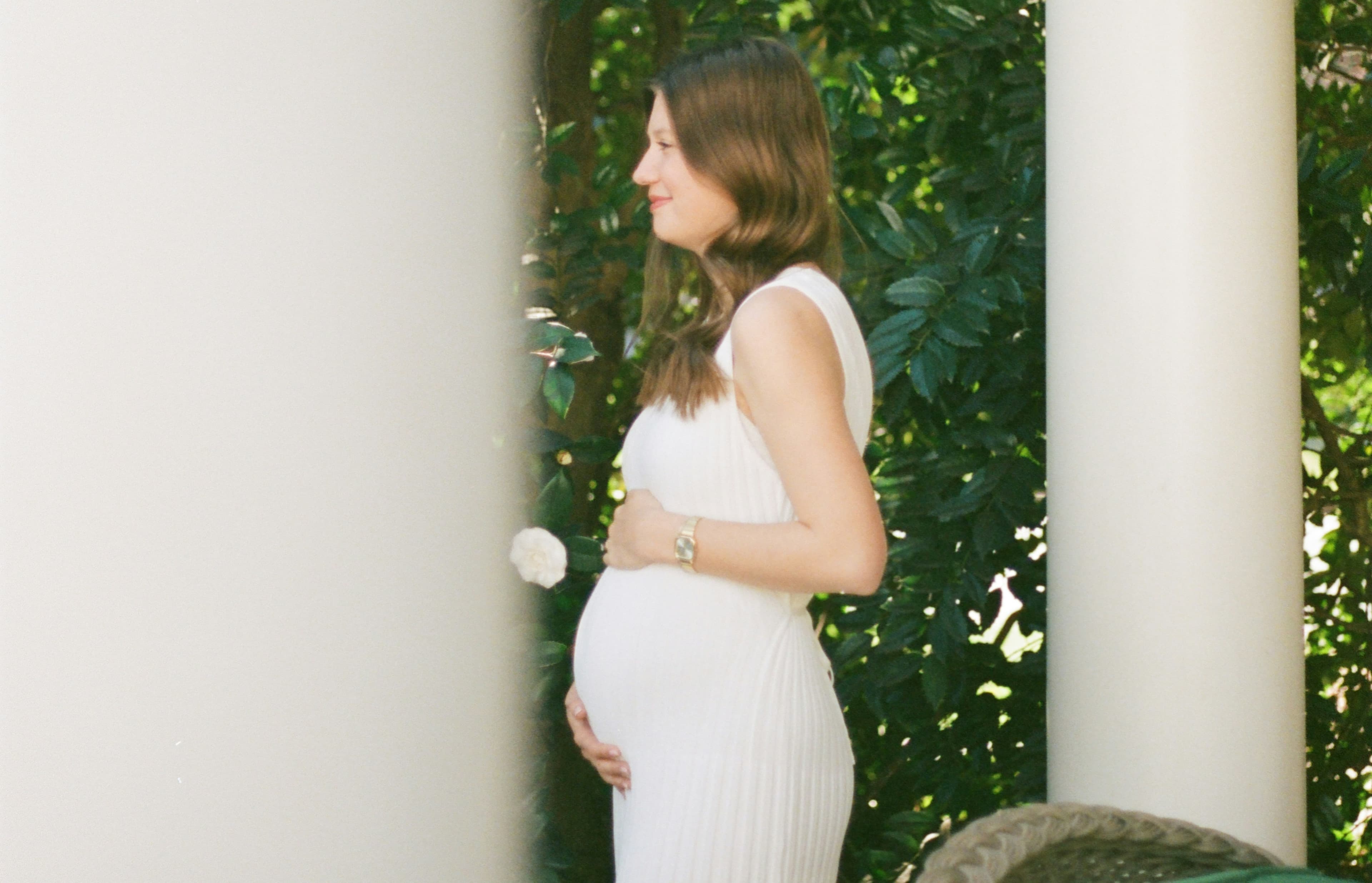 Pregnant woman in a white dress stands outdoors, holding her belly, surrounded by greenery and white columns.