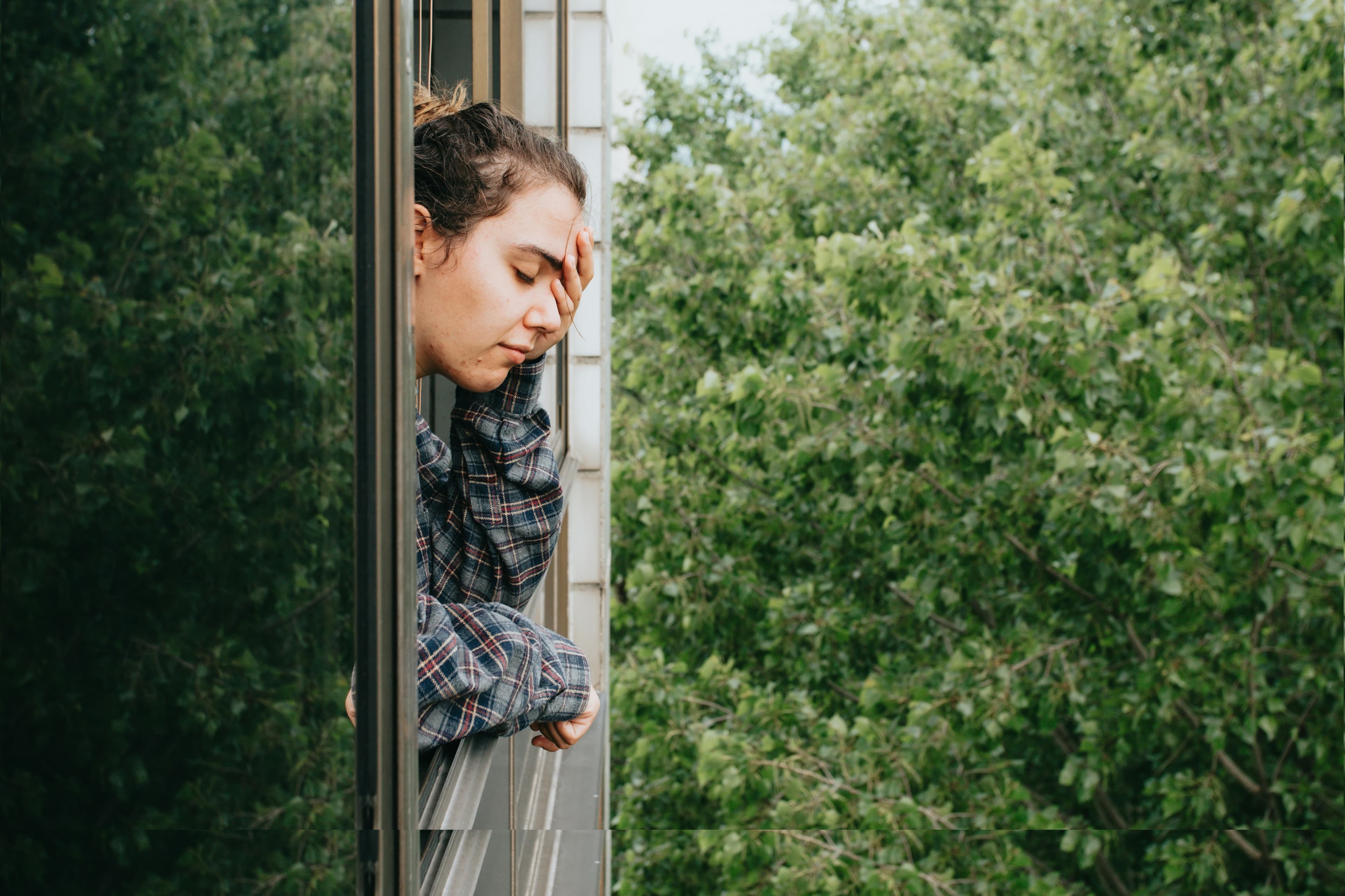 Person in a plaid shirt leans out of a window, resting their head on one hand, with a backdrop of lush green trees.